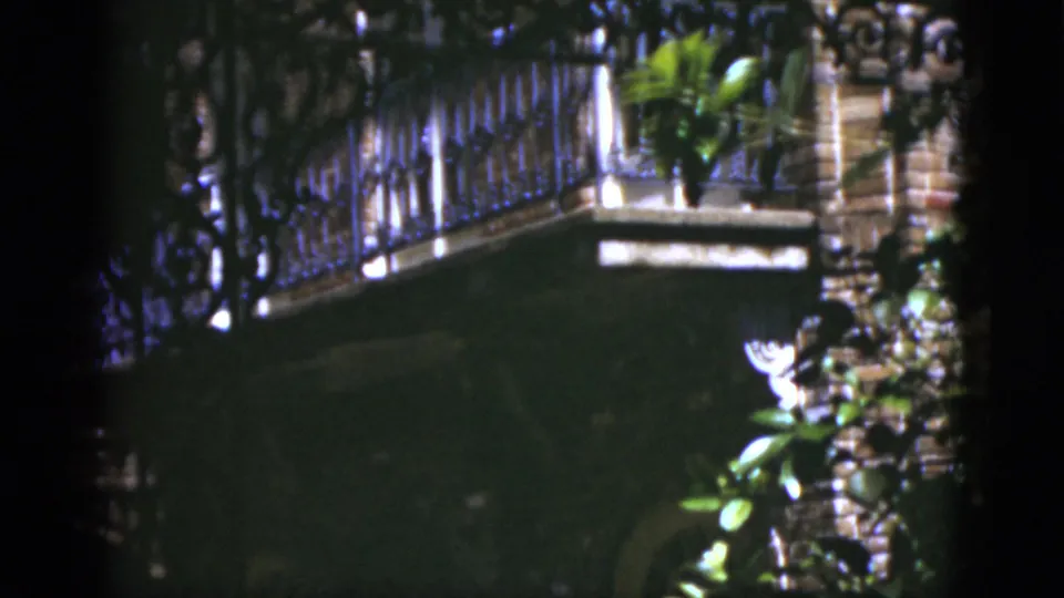 1975 Florida 8mm frame with a wrought-iron balcony railing, plants, white flowers, and a shaded exterior opening.