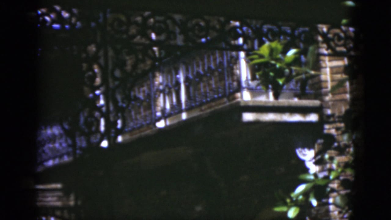 Ornate iron balcony railing beside brickwork and greenery in bright sunlight.