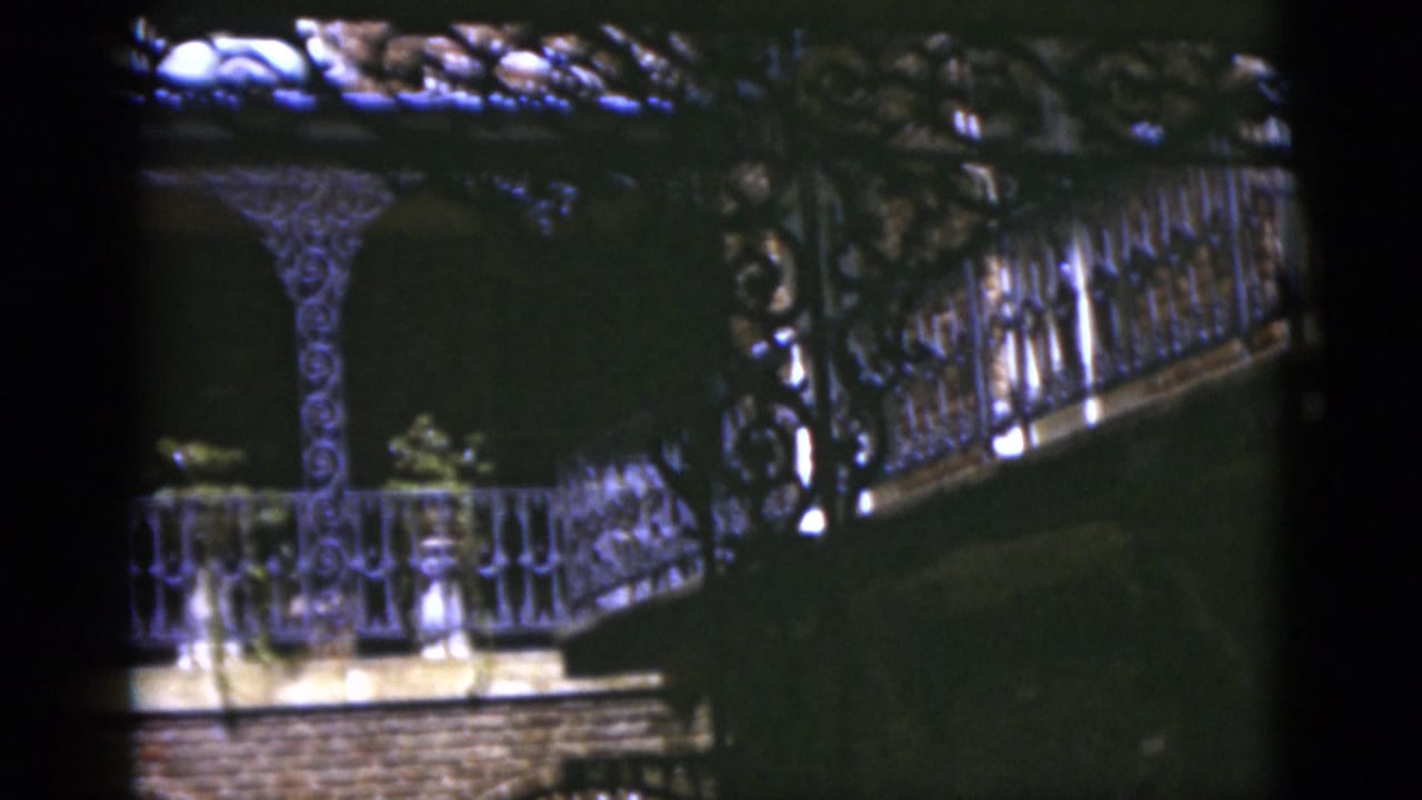 1975 Florida 8mm frame of an ornate exterior balcony with iron railings, decorative posts, brickwork, and dark arch openings.