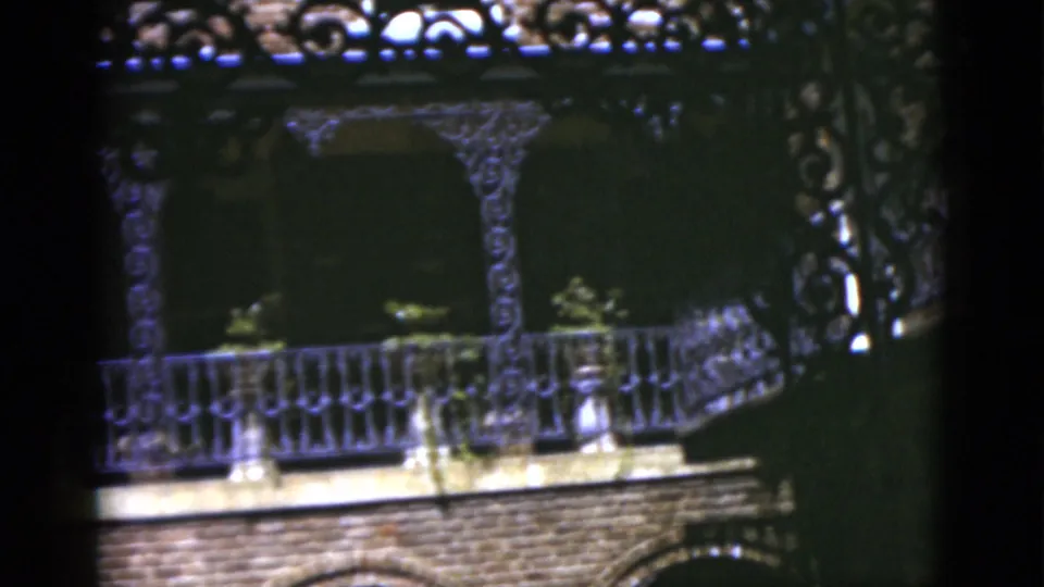 1975 Florida 8mm frame with wrought-iron balcony detail, brick base, potted greenery, and strong sun-shadow contrast.