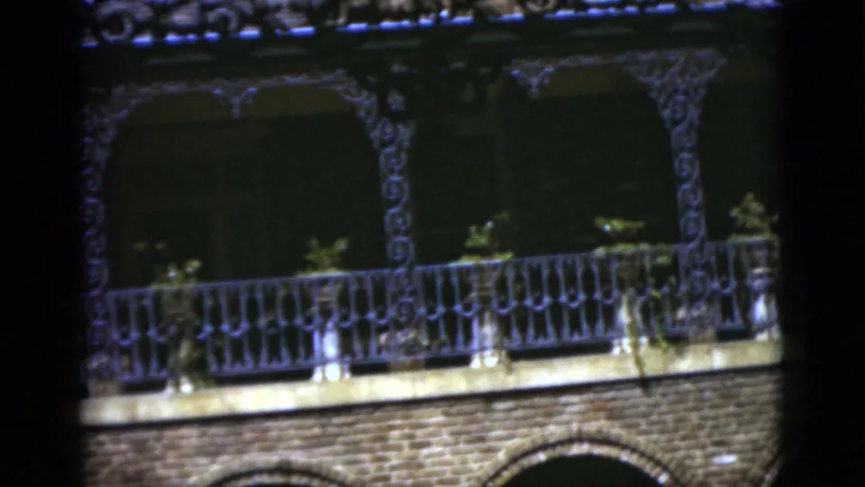 1975 Florida 8mm frame showing a wider view of ornate balcony railings, brick facade, shadowed arches, and small plants.