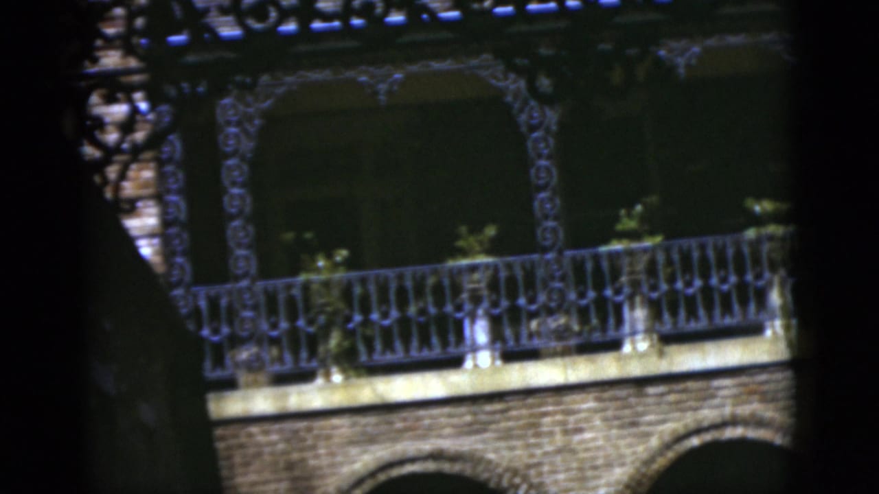 Brick house facade with ornate balcony railing partly hidden by tree shadows.