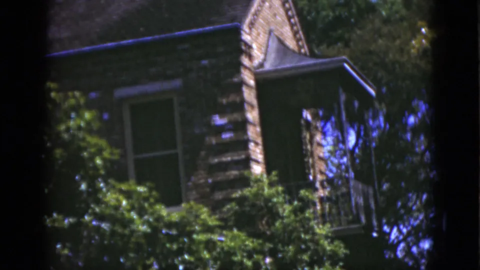 The camera holds on the upper red brick facade, with a window, roofline, and trees framing the house exterior.