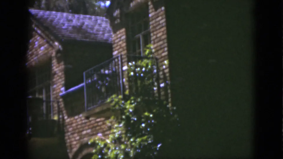 1975 Florida home-movie frame showing a brick balcony, metal railing, window, and thick greenery along the lower wall.