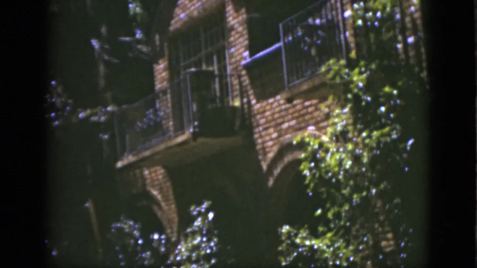 1975 Florida 8mm frame showing a brick exterior, balcony railing, arched support, and sunlit foliage.