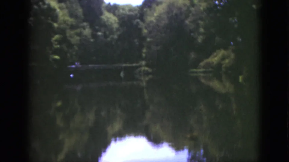 1951 Florida home-movie frame showing a quiet pond, tree reflections, shaded banks, and a small distant bridge.