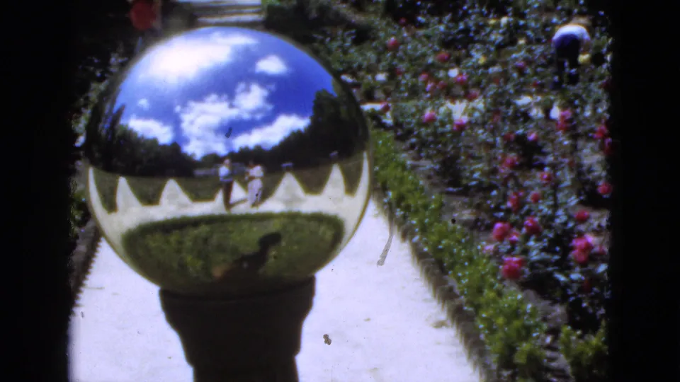 1951 Florida 8mm frame showing a polished garden sphere reflecting clouds, lawn, path, and faint adult figures in a flower garden.