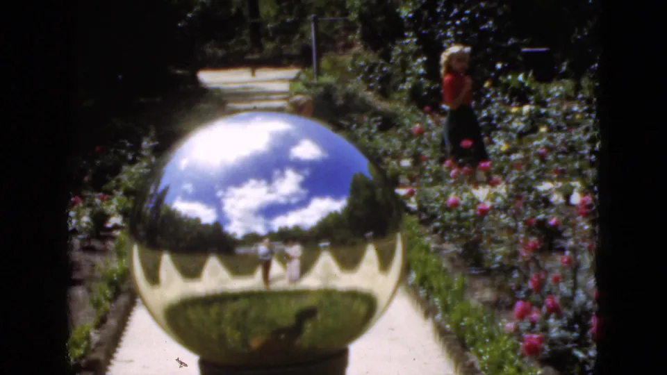 1951 Florida home-movie still showing a mirrored garden ball, rose beds, walkway, and an adult woman standing in the background.
