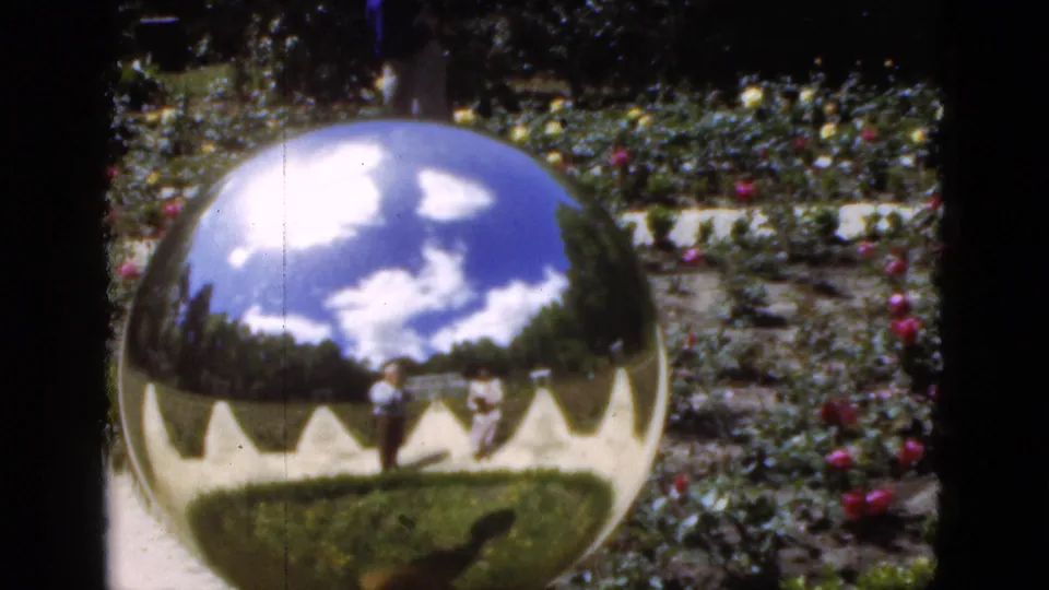 1951 Florida 8mm frame with a polished garden sphere reflecting clouds, lawn, path, and adult visitors in a landscaped flower garden.