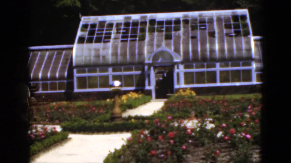 A glass greenhouse in the Florida garden appears behind flower beds, a paved approach, trimmed plantings, and a small lamp fixture.
