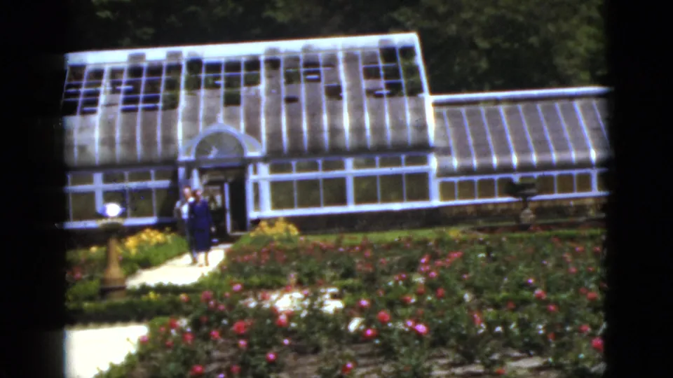Adult visitors walk along a path before the white greenhouse and flowering beds in this 1951 Florida garden scene.