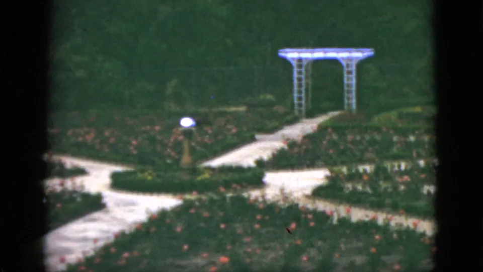 1951 Florida 8mm frame of formal garden beds, curving pale walkways, lawn, and a white arbor in soft focus.