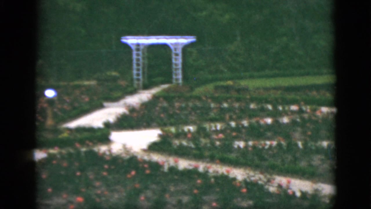Garden paths and flower beds with a white arbor in the distance.