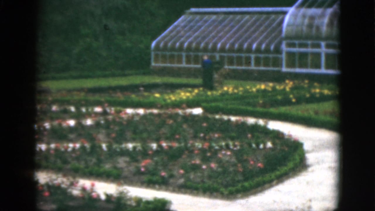 Greenhouse beyond curved garden paths and flower beds with a distant visitor standing on the lawn.
