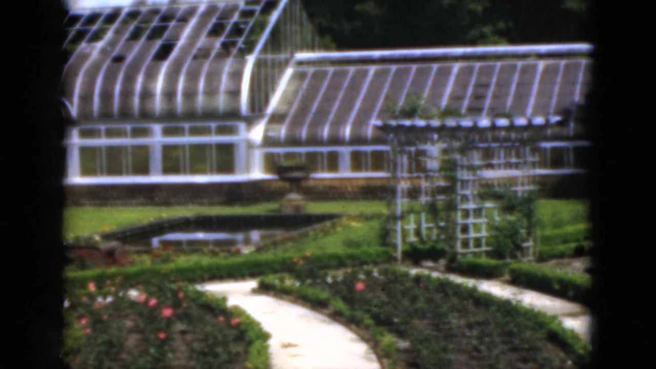 Greenhouse and trellis beside a formal garden with paths, hedges, flowers, and a pond.
