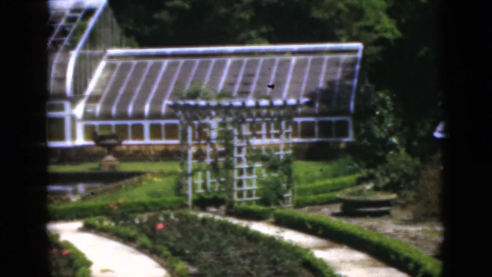 1951 Florida 8mm still focused on greenhouse windows, white trelliswork, formal plant beds, a pond edge, and a curving walk.
