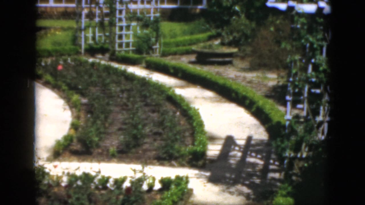 1951 Florida garden view with pale walkways, low hedges, rose planting beds, white trellises, and a small gate or bench form.