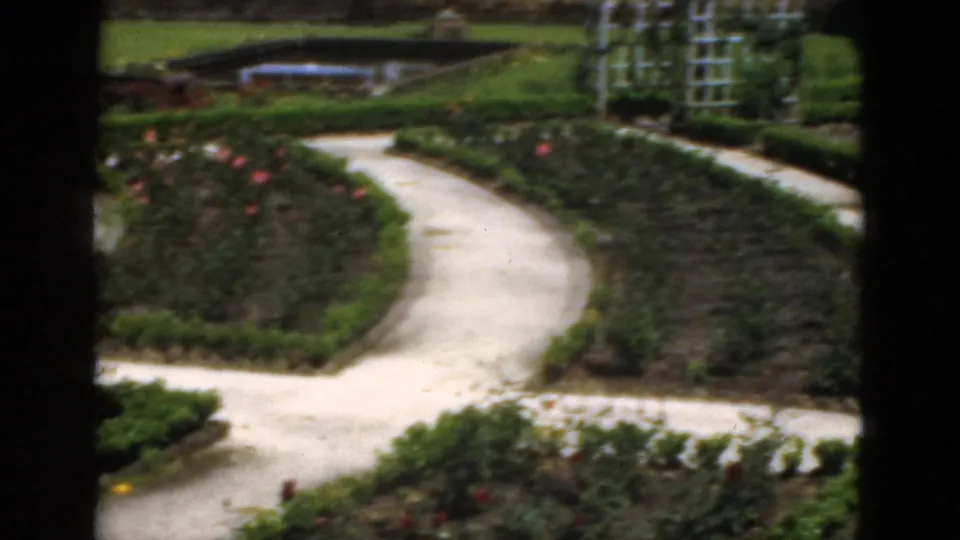 1951 Florida 8mm frame showing intersecting garden paths, clipped borders, rose beds, white trellises, lawn, and a small pond in the background.