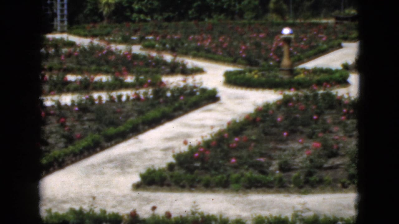 Formal rose garden with pale gravel paths, planting beds, and a small ornamental lamp.