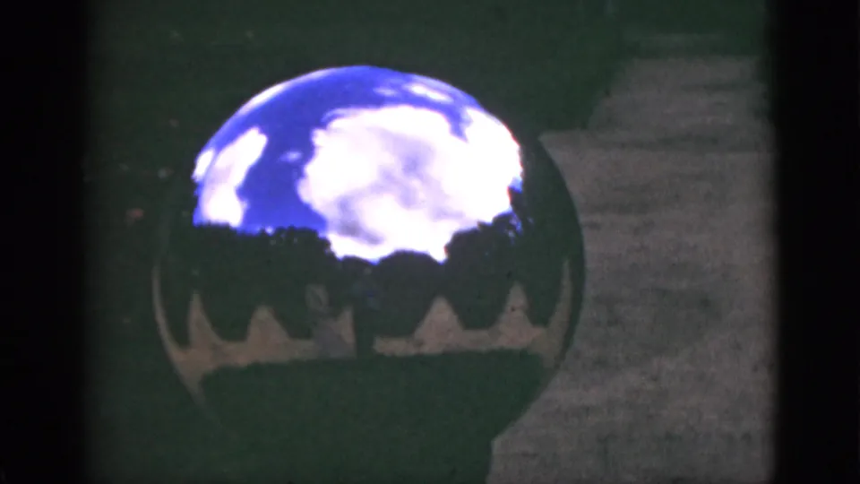1951 Florida garden detail with a reflective globe ornament beside a walkway, mirroring blue sky, white clouds, and tree silhouettes.