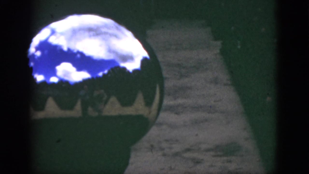 1951 Florida 8mm frame showing a shiny garden sphere beside a path, reflecting blue sky, clouds, and silhouetted foliage.