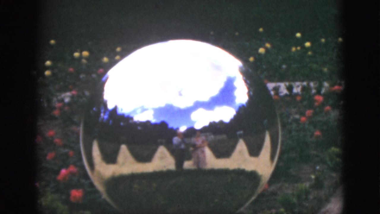 Reflective garden sphere with clouds, flowers, scalloped base, and faint figures in the reflection.