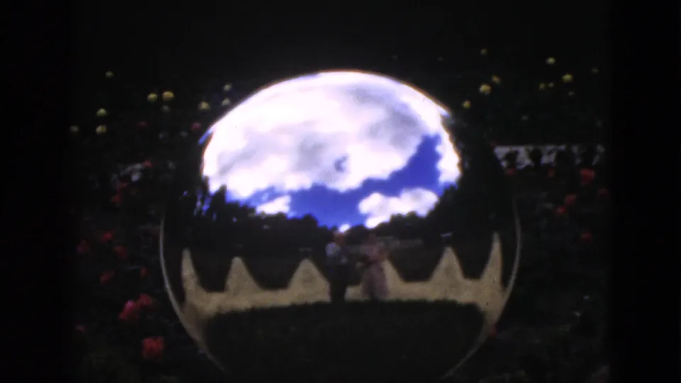 1951 Florida 8mm frame with a shiny garden sphere reflecting clouded sky, dark foliage, red flowers, and two faint people.