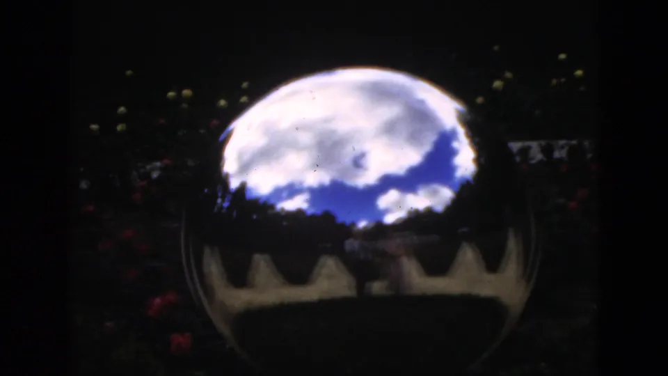 1951 Florida home-movie frame of a reflective garden ball with clouded sky, silhouetted trees, red flowers, and darkened film edges.