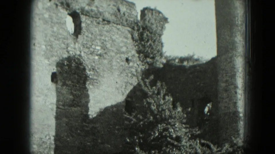 1951 West Germany home-movie frame of ruined stonework, a tower-like wall opening, and dense plants at the base.