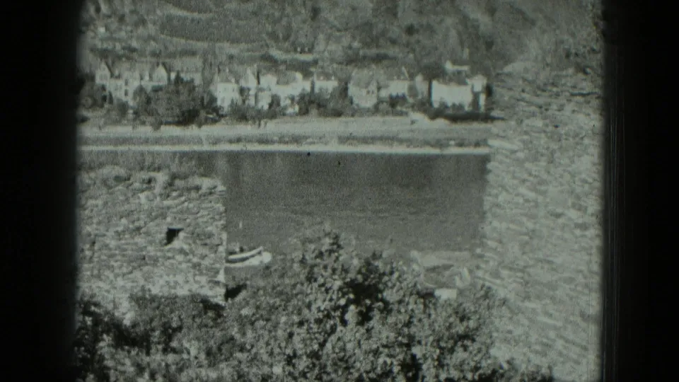 1951 West Germany 8mm frame looking past castle stones to a river, small boat, and white houses on the far bank.