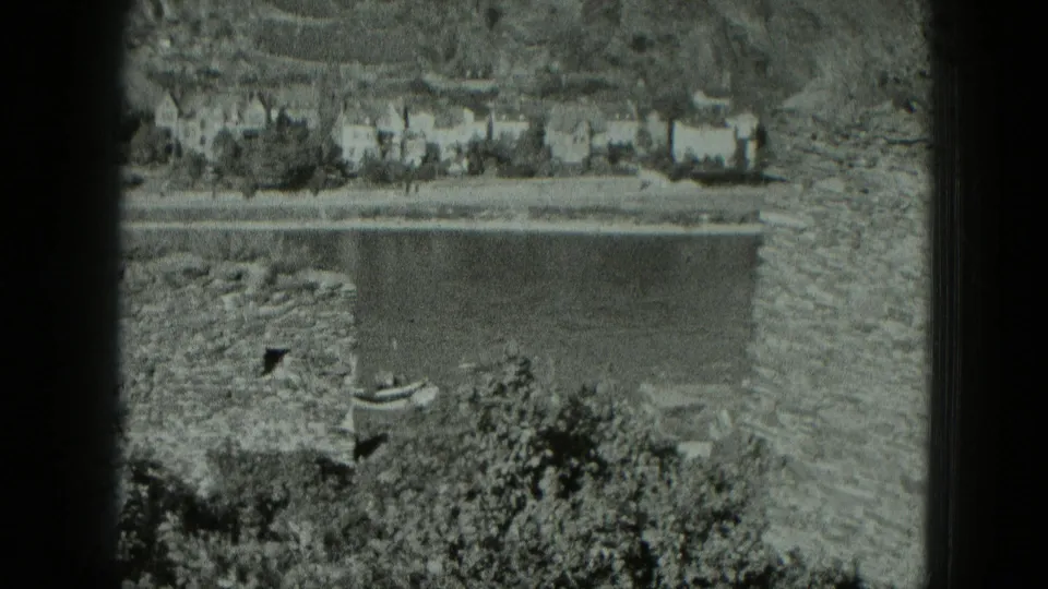 1951 West Germany frame showing a river settlement and small boat beyond foreground stone ruins and vegetation.