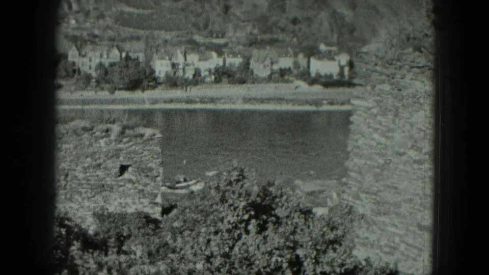 1951 West Germany 8mm frame showing a rough stone wall, a dark opening, nearby vegetation, and possible water or shoreline in the background.