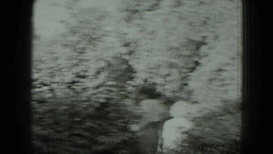 Blurred black-and-white 8mm frame, likely from handheld camera movement near the ruin site, with soft mottled ground or stone texture.