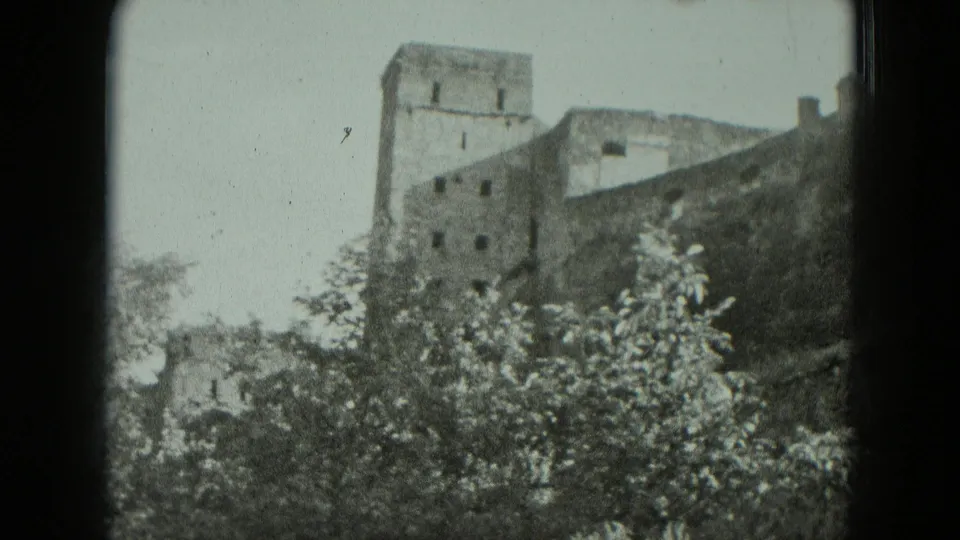1951 Germany 8mm frame showing the castle ruin from below, with a tower, wall sections, and thick trees in front.