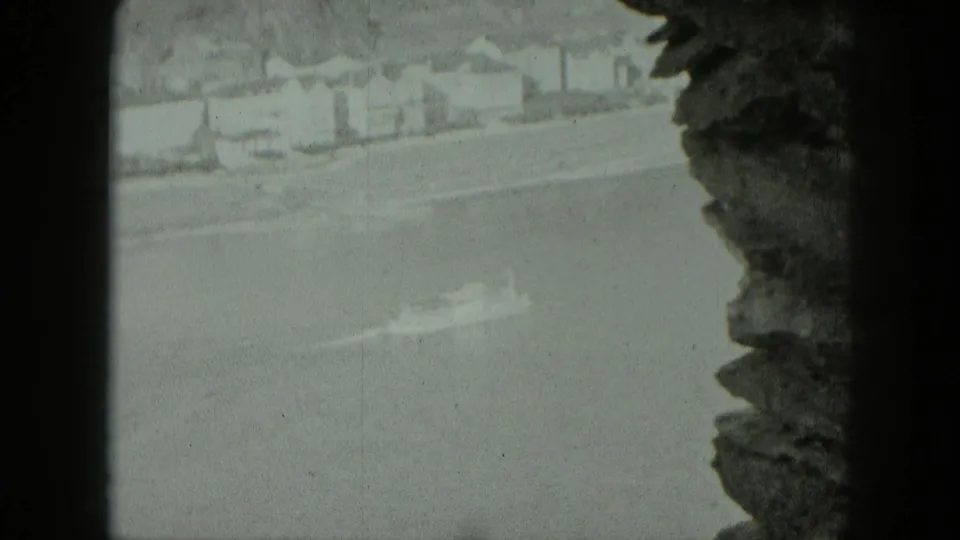A river or road with pale buildings is viewed through a stone window-like opening in the supplied 1951 Germany home-movie footage.