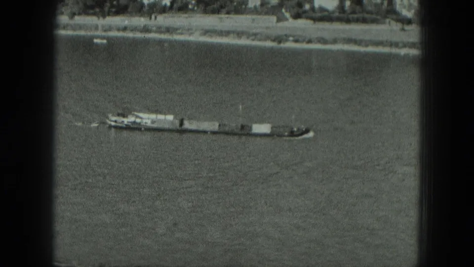 A long river vessel moves across a 1951 West Germany waterway, with a stone bank and shoreline buildings behind it.