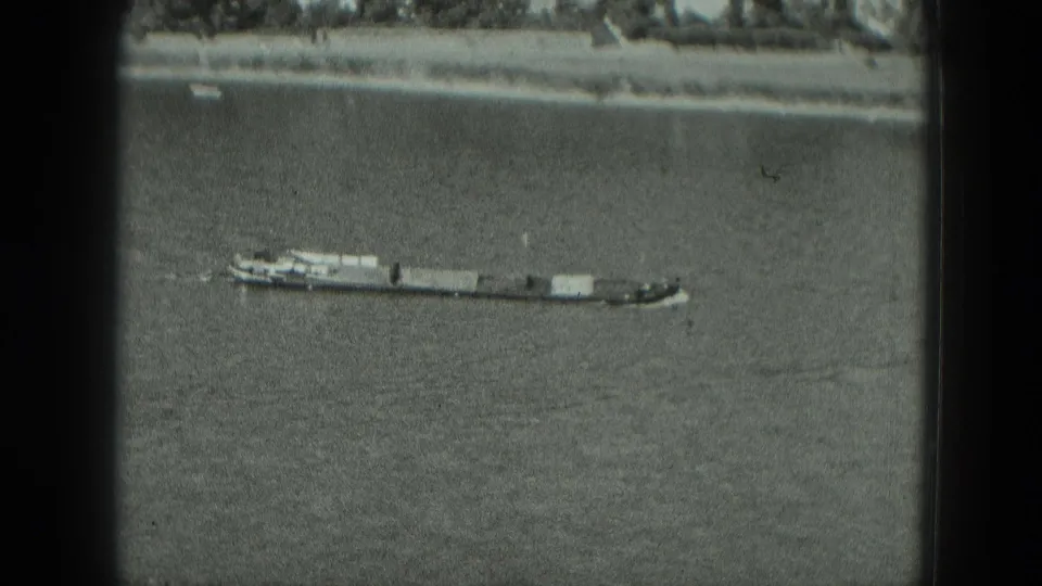 Elevated 1951 Germany 8mm view of a tugboat pushing barges on a broad river, with trees and buildings along the far bank.