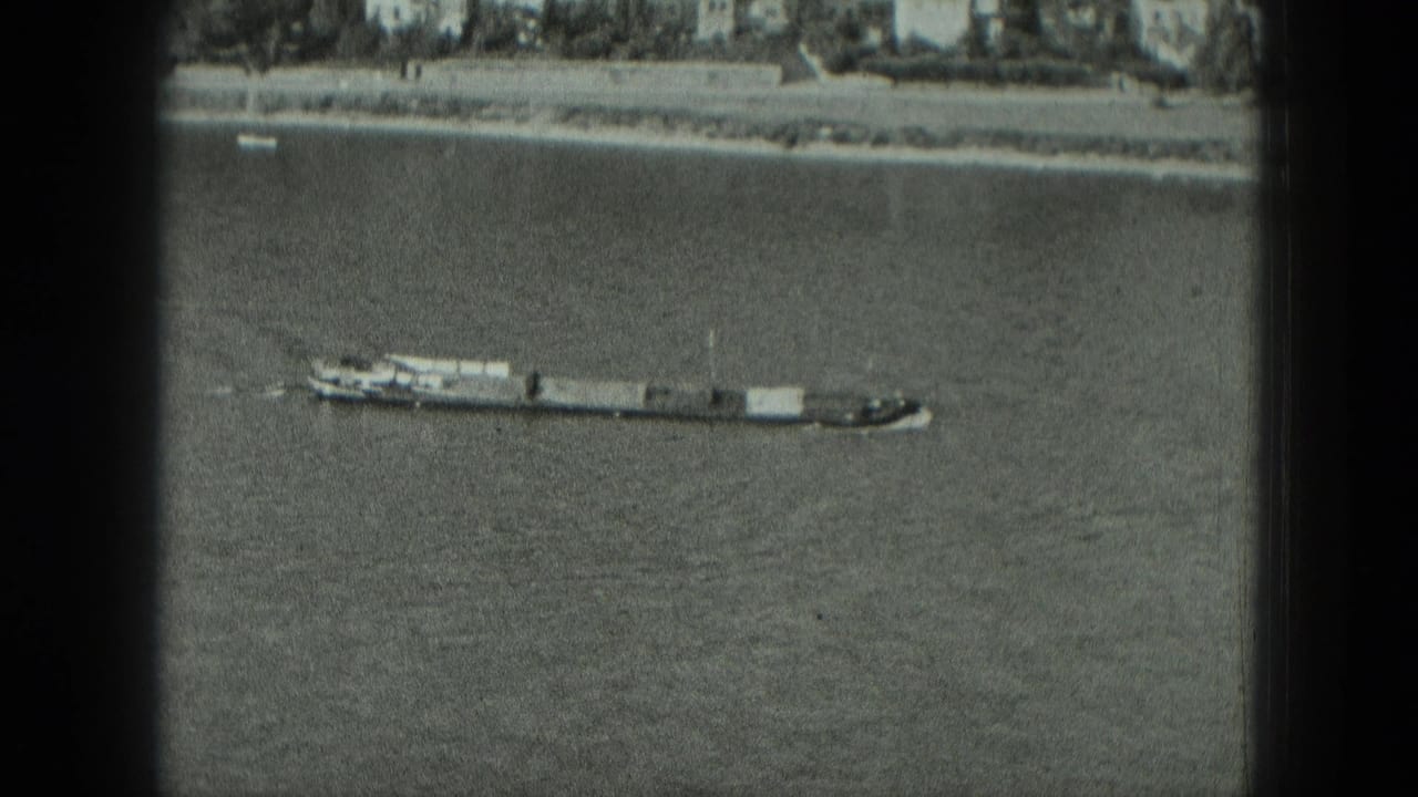 Long barge tow on river water below a distant embankment and buildings.