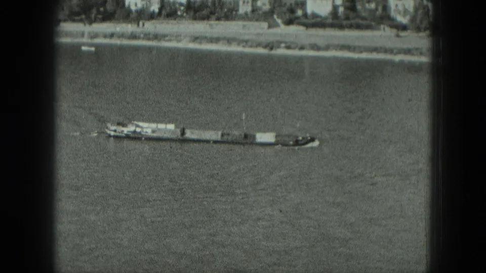 Black-and-white elevated view of a tug and barges on a German river, framed by visible 8mm film borders and a distant built shoreline.