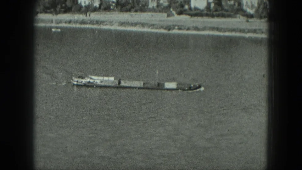 1951 8mm home-movie frame showing a tugboat pushing barges upstream on a wide river in Germany, with the far bank visible above.