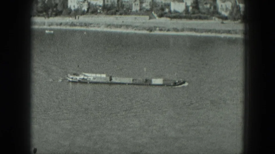 Elevated black-and-white 8mm view of a tugboat pushing barges on a West German river, with a distant shoreline and scanned frame edges.