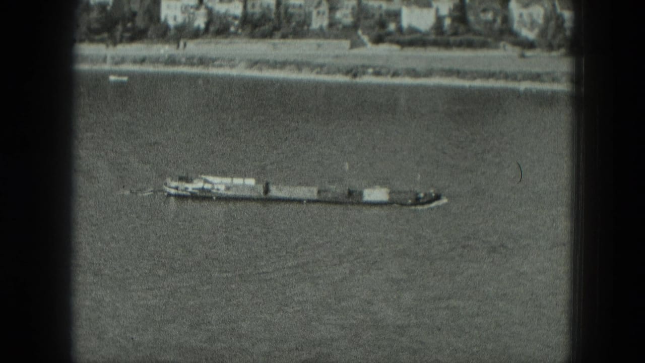 1951 8mm frame of a long barge moving across a wide river in Germany, with a distant bank and buildings beyond.