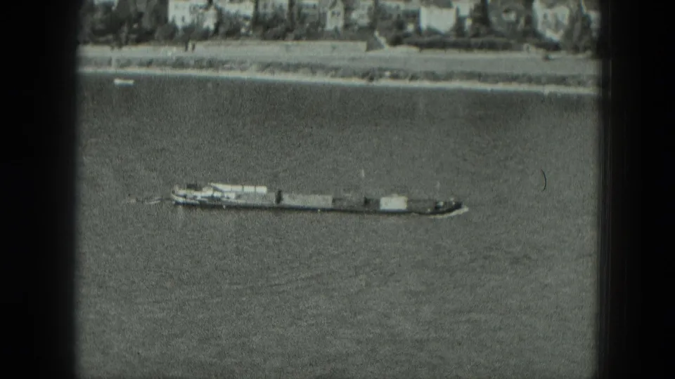 1951 8mm frame of a long barge moving across a wide river in Germany, with a distant bank and buildings beyond.
