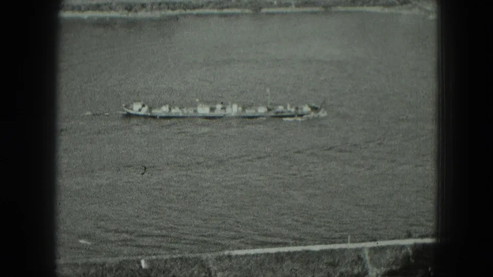 1951 Germany 8mm frame showing a long vessel crossing calm water near a low shoreline and embankment.