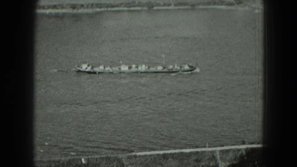 1951 Germany 8mm frame showing a long cargo boat moving across calm water above a waterfront embankment.
