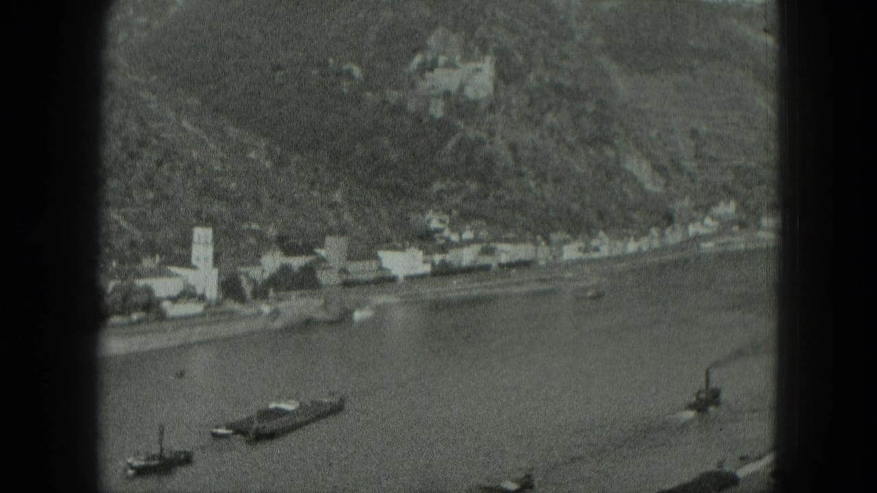 Riverside town below a wooded hillside with boats on the river and a church tower near the bank.