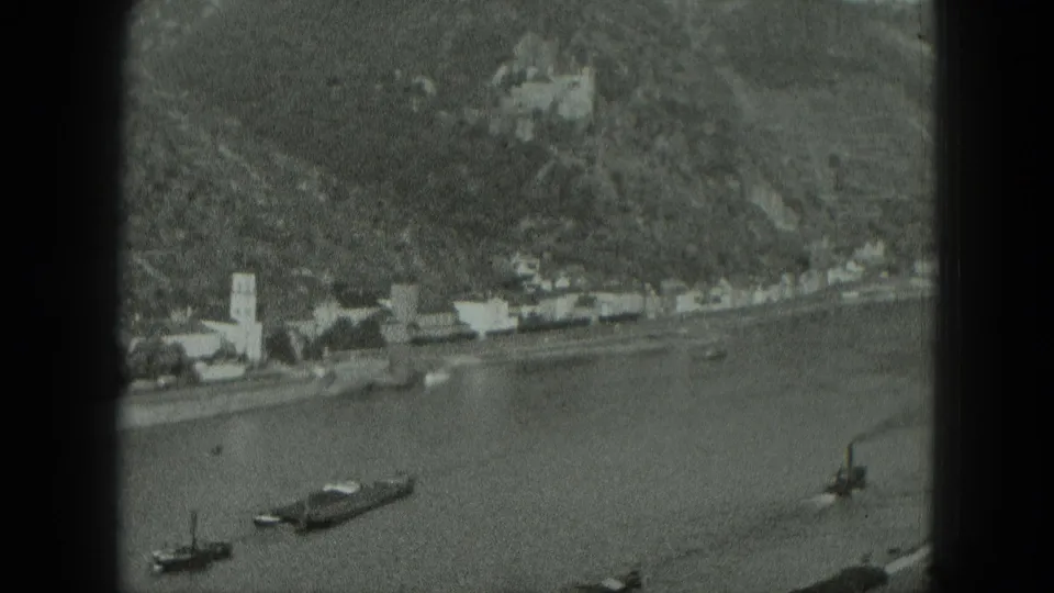 1951 West Germany 8mm frame showing a river town, church steeple, boats, and a hilltop castle-like structure in the distance.