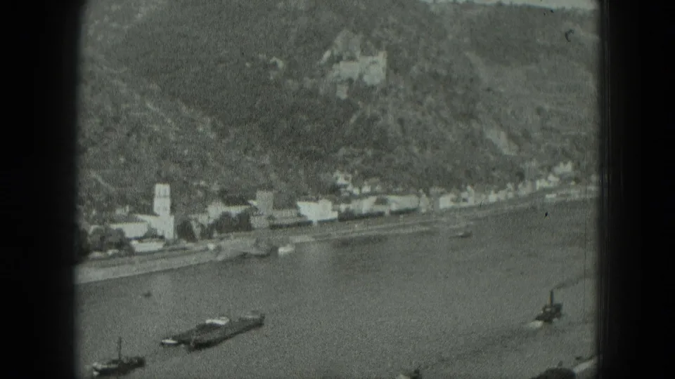 Black-and-white 8mm view of a West German riverside settlement with boats, a church tower, and hillside architecture.
