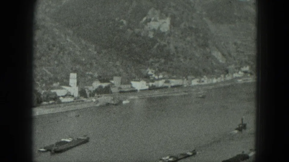 1951 black-and-white home-movie frame of a German riverbank town with boats, a church steeple, and steep wooded terrain.