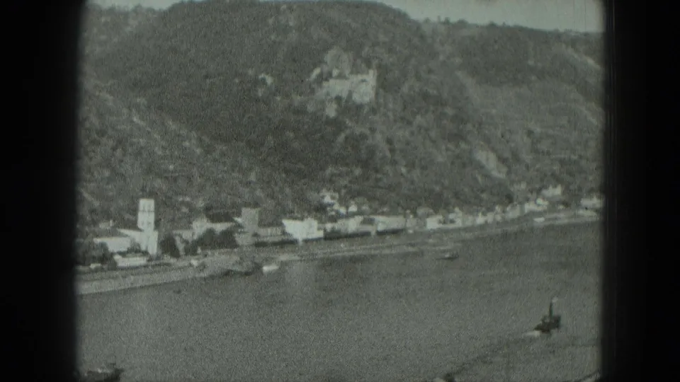 West Germany 1951 8mm frame showing a river town beneath a wooded hill with boats, a church tower, and a distant castle-like building.